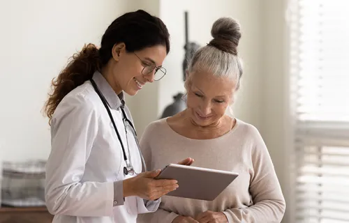 Happy female doctor and senior patient look at tablet screen discuss treatment or therapy on gadget.